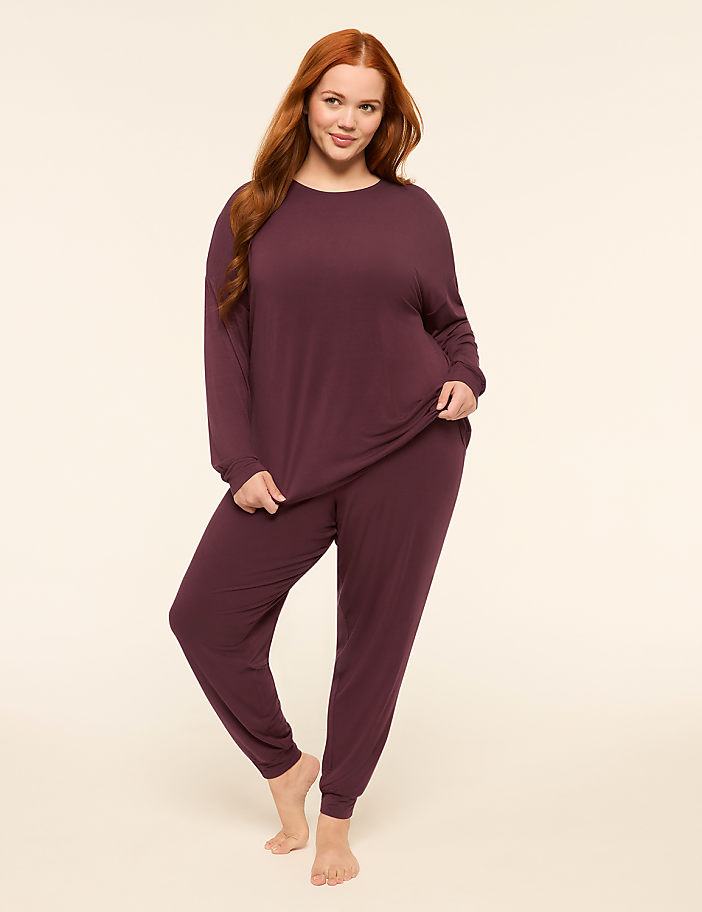 A woman with long red hair stands in a plum-colored pajama set against a plain, light-colored background.