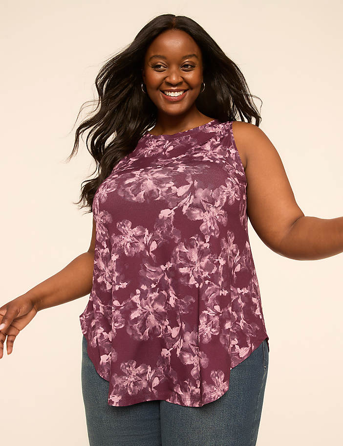 A smiling woman in a floral top and jeans stands against a plain, light-colored background.