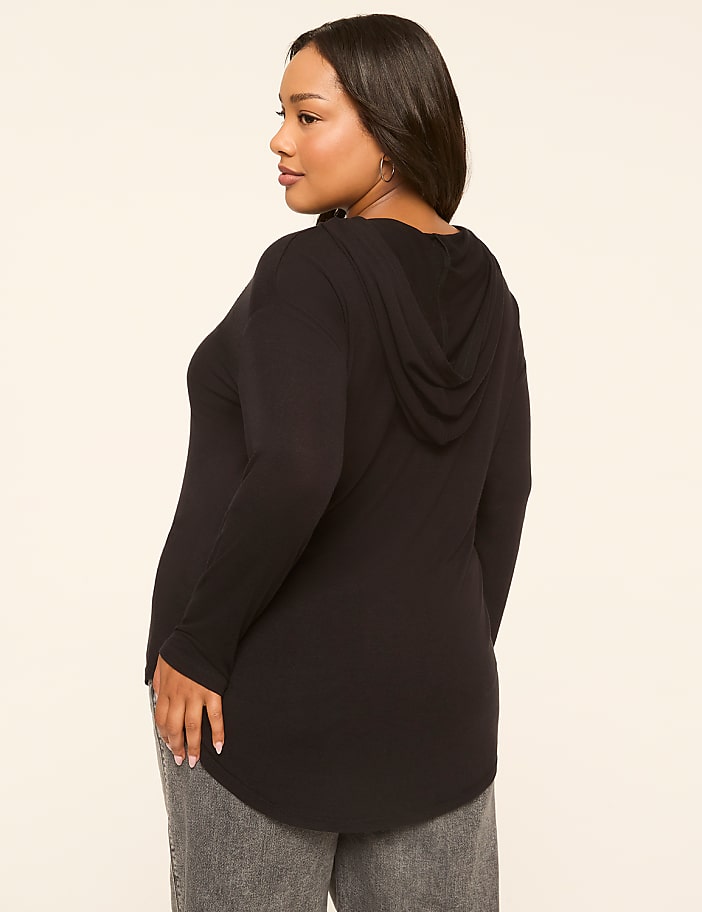 A woman in a black hooded shirt and gray jeans stands against a plain, light-colored background.
