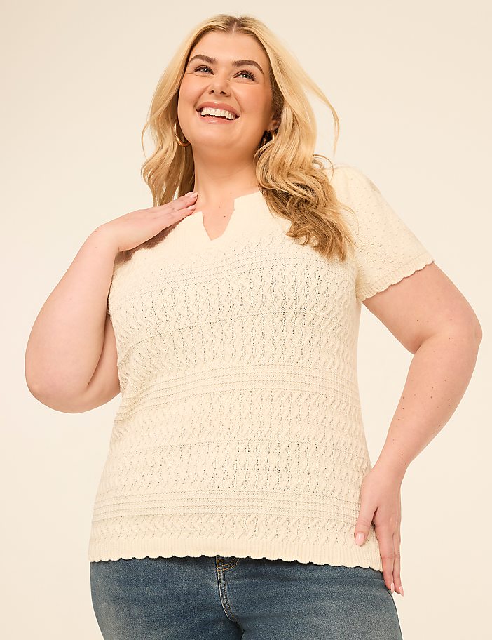 A smiling woman in a cream-colored, textured knit top and blue jeans poses against a plain, light-colored background.
