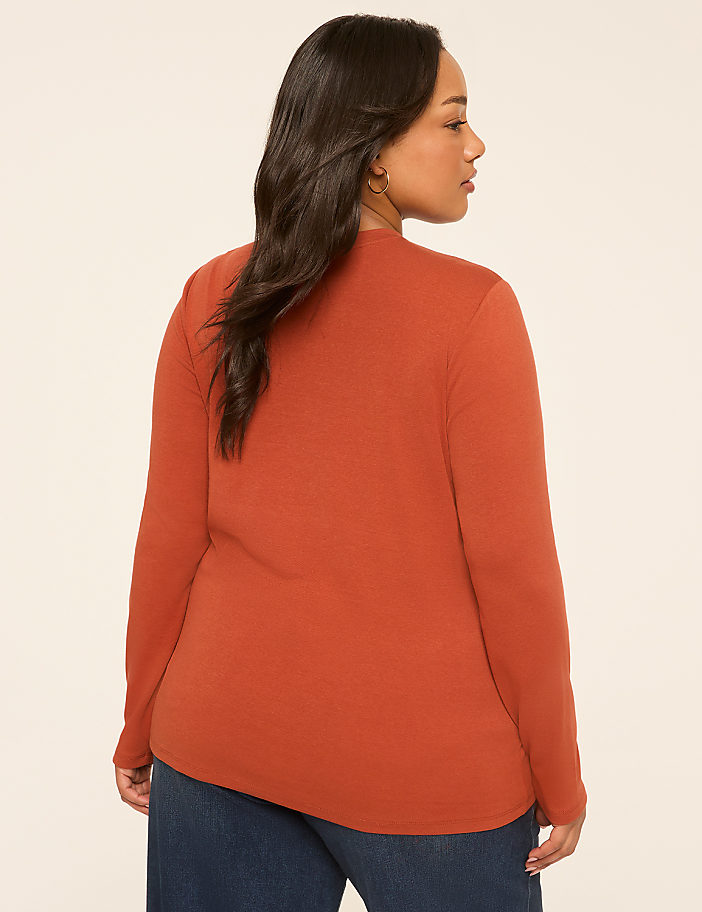 A woman in a rust-colored long-sleeved shirt and blue jeans stands against a plain, light-colored background.