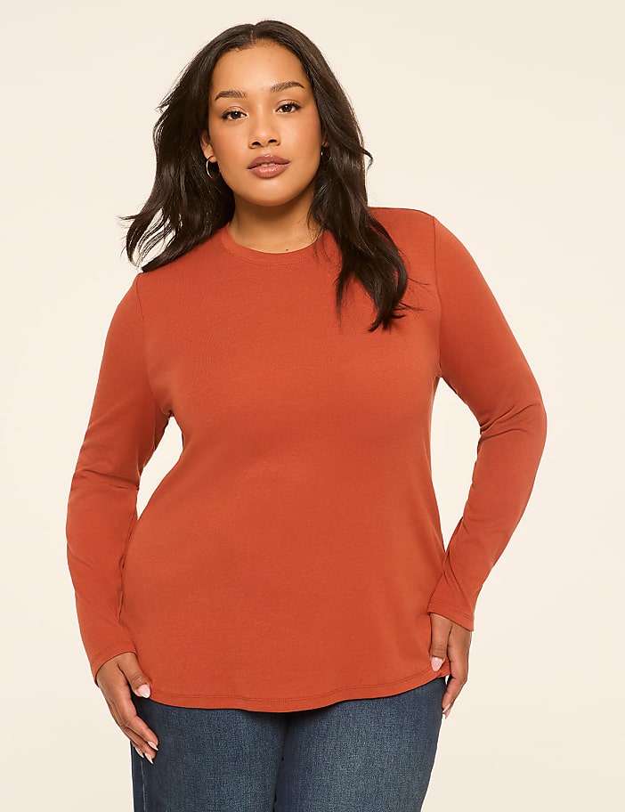 A woman in a rust-colored long-sleeved shirt and blue jeans stands against a plain, light-colored background.