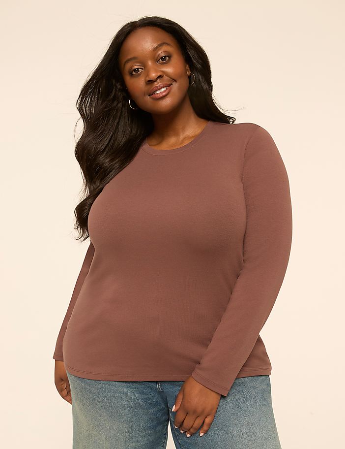 A smiling woman with long dark hair wears a brown long-sleeved shirt and blue jeans against a plain, light-colored background.