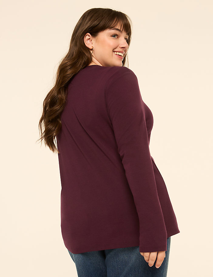 A woman in a burgundy long-sleeved shirt and blue jeans smiles over her shoulder against a plain, light-colored background.