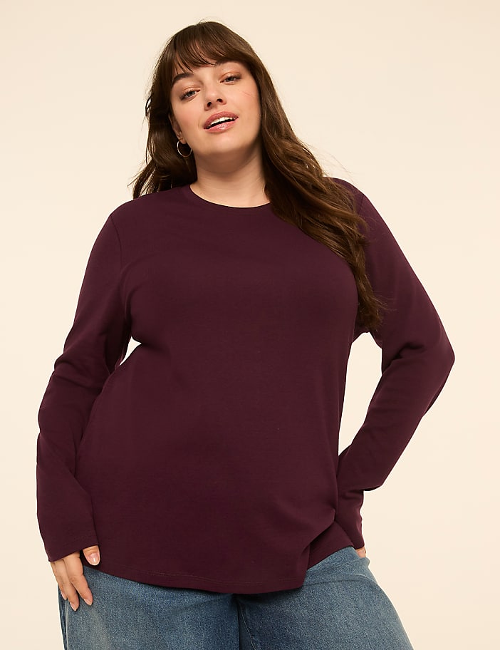 A woman in a burgundy long-sleeved shirt and blue jeans stands against a plain, light-colored background.