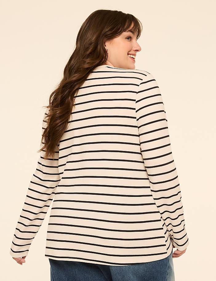 A woman with long brown hair smiles over her shoulder, wearing a cream and black striped long-sleeved shirt against a plain beige background.