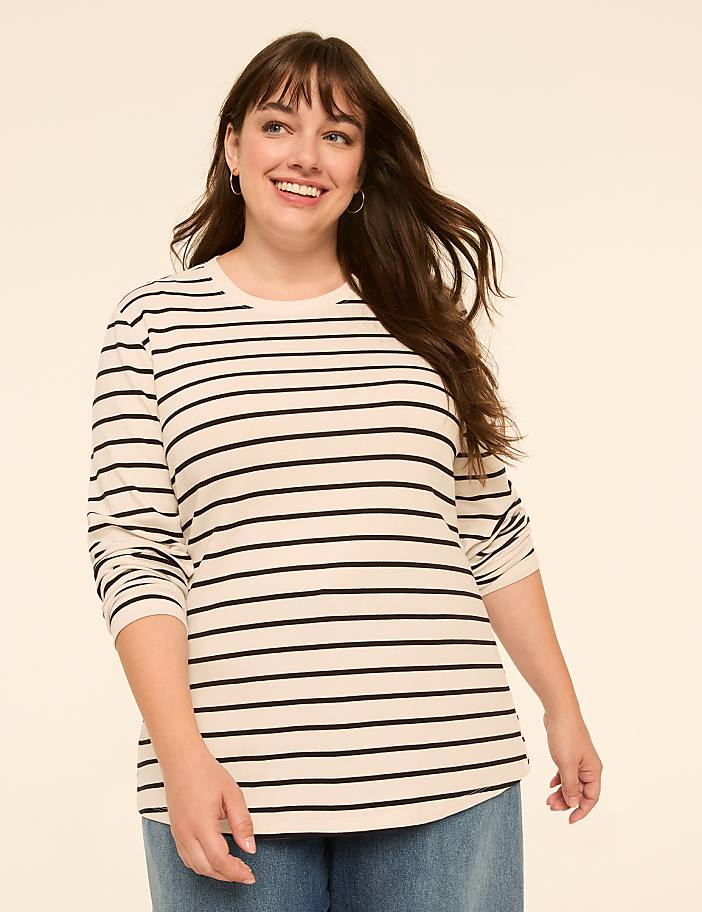 A smiling woman with long brown hair wears a striped shirt and jeans against a plain, light-colored background.