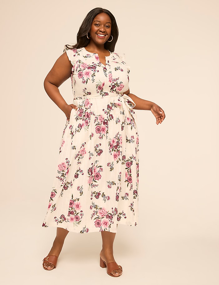 A smiling woman in a floral dress stands against a plain, light-colored background.