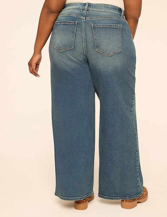 A person wearing wide-leg blue jeans and leopard print shoes stands against a plain, light-colored background.