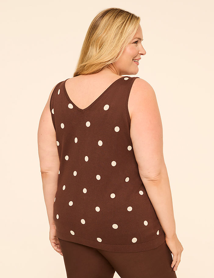 A woman in a brown polka dot top and brown pants stands against a plain cream background.