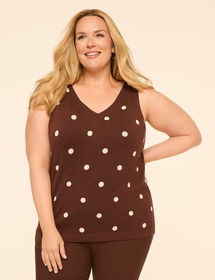 A smiling woman in a brown polka-dot tank top and brown pants stands against a plain, light brown background.