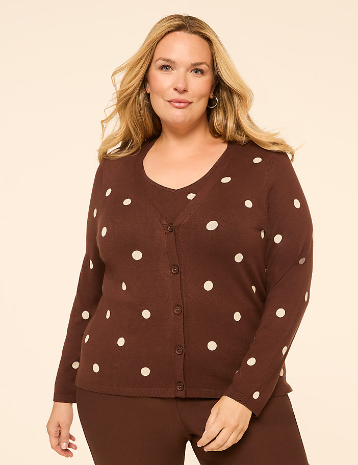 A woman in a brown polka dot cardigan and matching pants stands against a plain, light brown background.