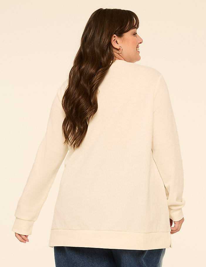 A woman with long, dark hair wearing a cream-colored sweatshirt and blue jeans stands against a plain, light-colored background.