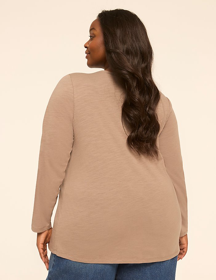 A woman in a tan long-sleeved shirt and blue jeans stands against a plain, light-colored background.