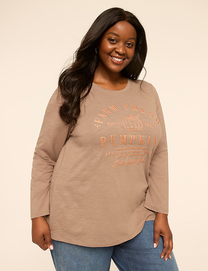 A smiling woman in a long-sleeved shirt and jeans stands against a plain, light-colored background.