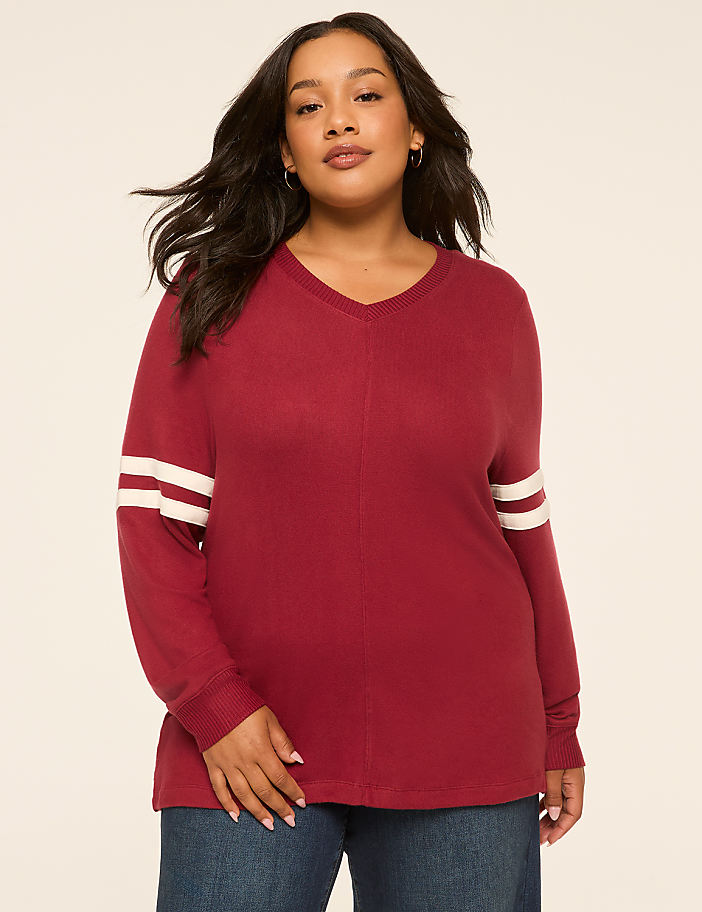 A woman in a maroon v-neck sweater with white stripes on the sleeves stands against a plain, light-colored background.