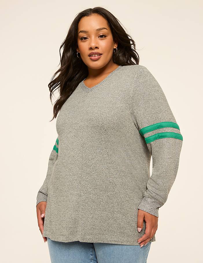 A woman in a grey v-neck sweater with green stripes on the sleeves stands against a plain beige background.