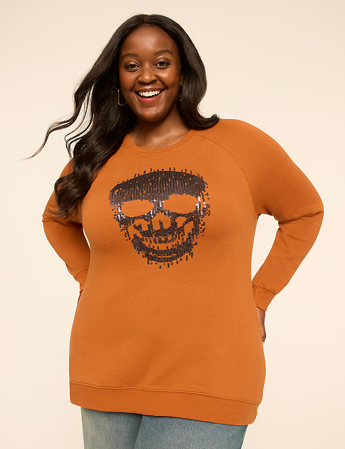 A smiling woman in a burnt orange sweatshirt with a sequined skull design stands against a plain, light background.