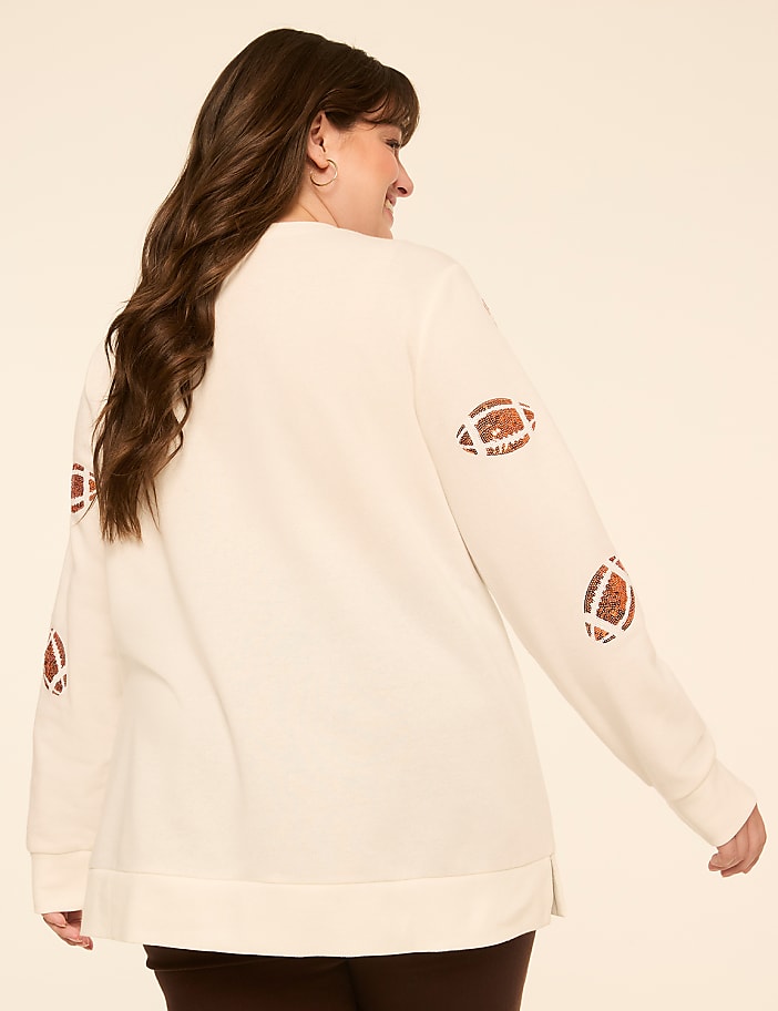 A woman in a cream-colored sweatshirt with football patches on the sleeves poses against a plain, light-colored background.