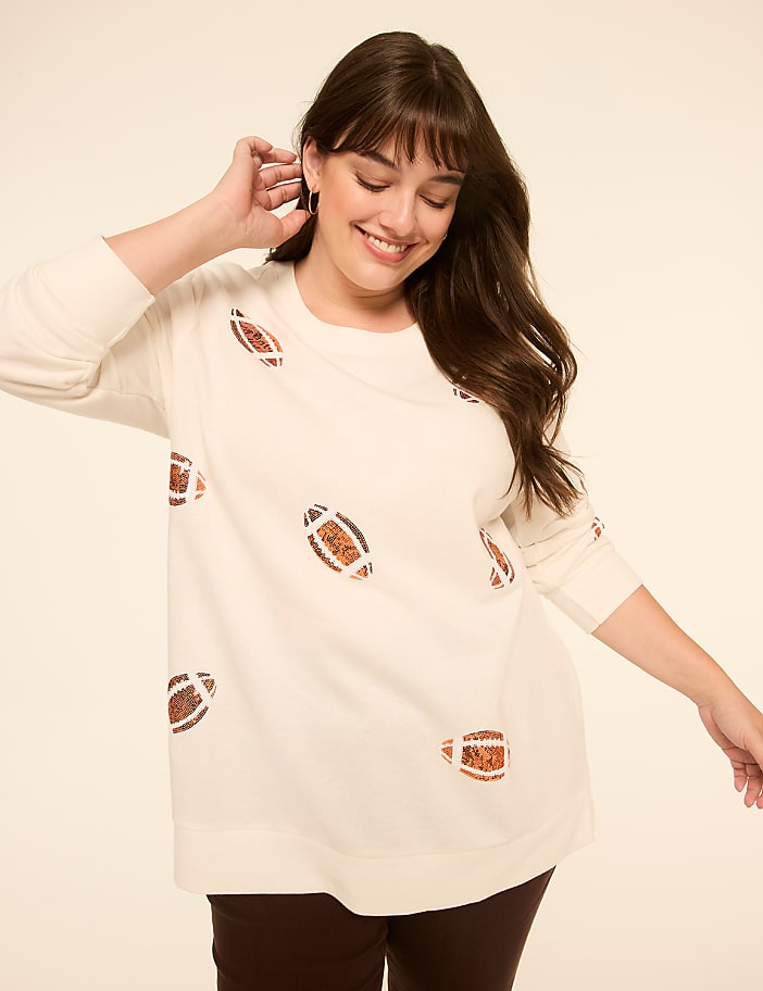 A smiling woman in a cream-colored sweater with football patterns stands against a plain, light-colored background.
