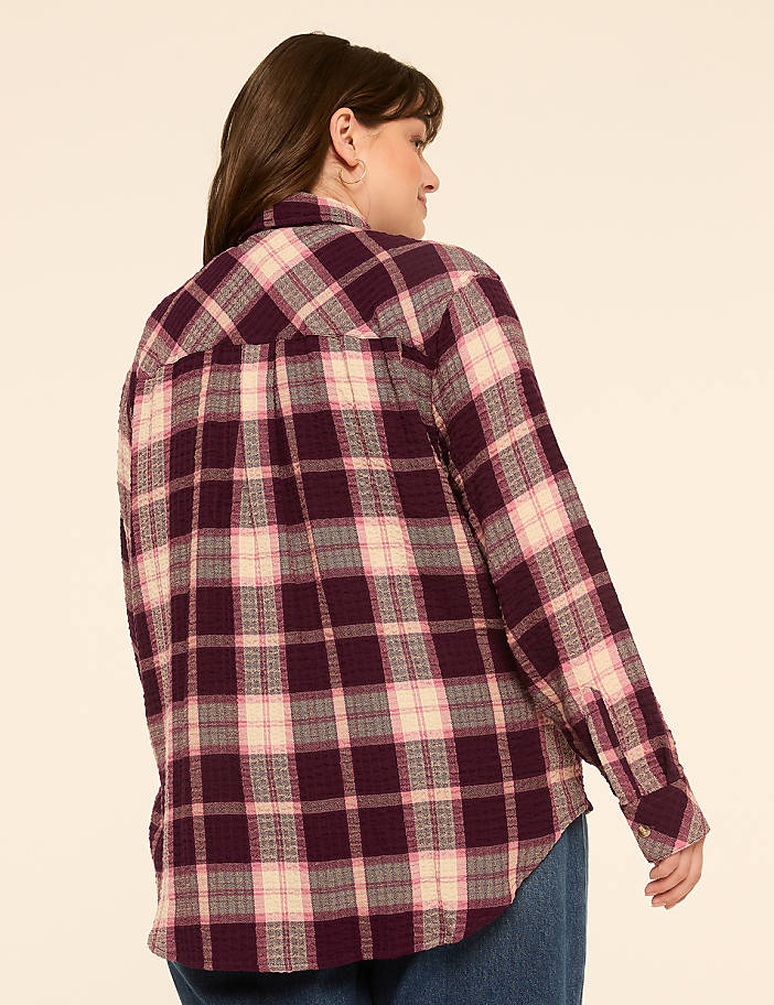 A woman in a purple plaid shirt stands against a plain, light-colored background.