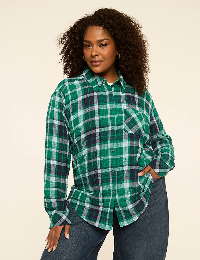 A woman in a green plaid shirt and blue jeans stands against a plain, light-colored background.