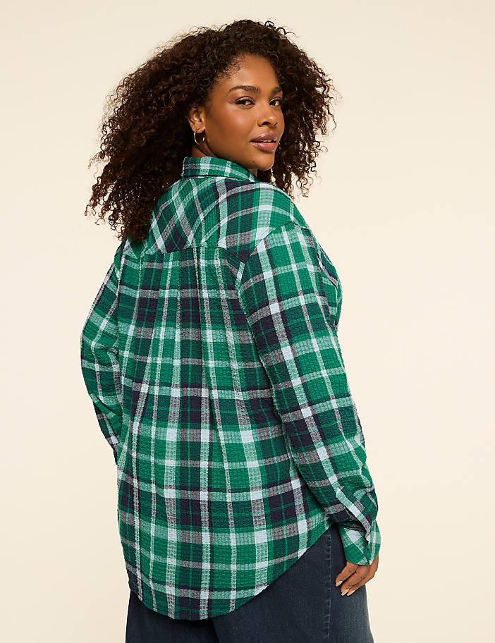 A woman with curly dark hair wearing a green plaid shirt stands against a plain, light-colored background.