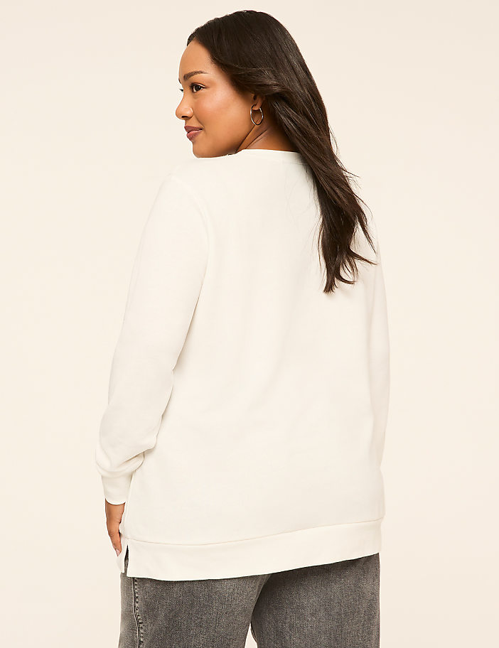 A woman in a white sweatshirt and grey jeans stands against a plain, light-colored background.