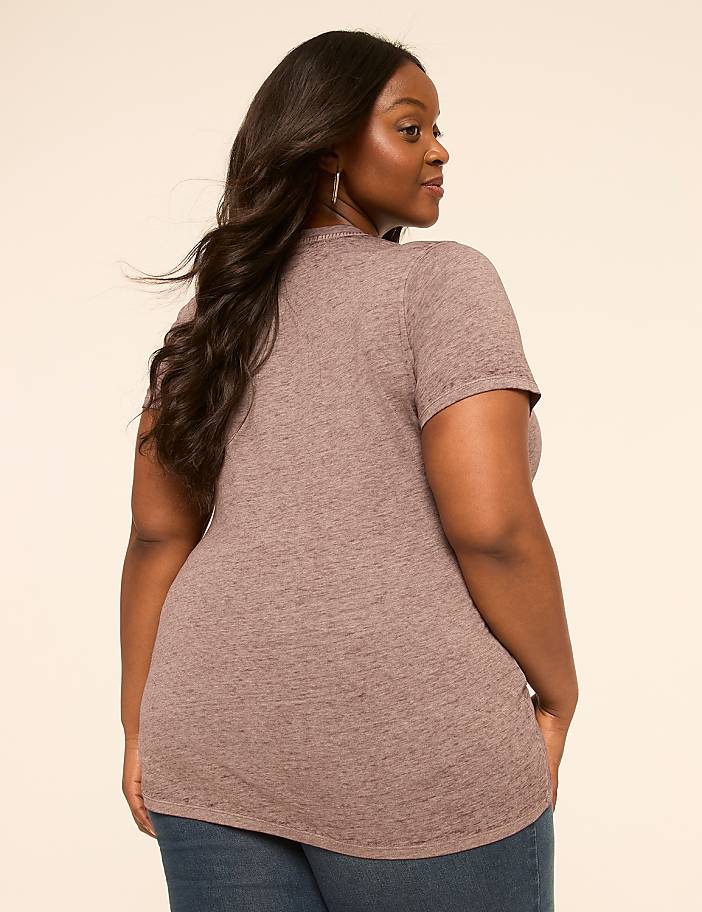 A woman in a heathered purple t-shirt and blue jeans stands against a plain, light-colored background.