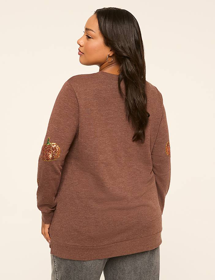 A woman in a brown sweater with sequined pumpkin patches on the elbows stands against a plain, light-colored background.