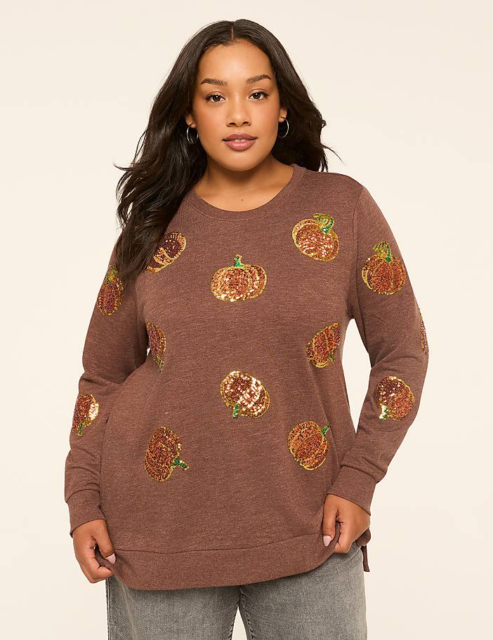 A woman in a brown sequined pumpkin sweatshirt stands against a plain, light-colored background.