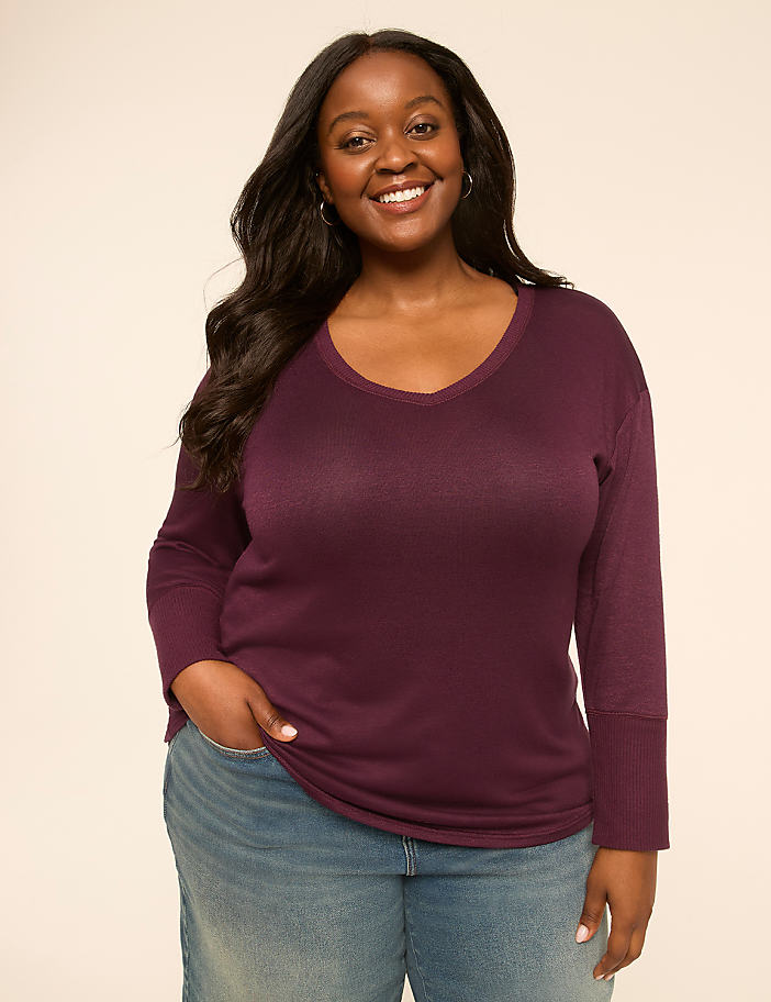 A smiling woman in a purple v-neck shirt and jeans stands against a plain, light-colored background.