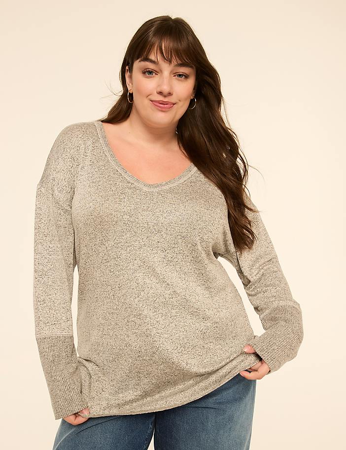 A woman in a gray v-neck sweater and blue jeans smiles against a plain, light-colored background.