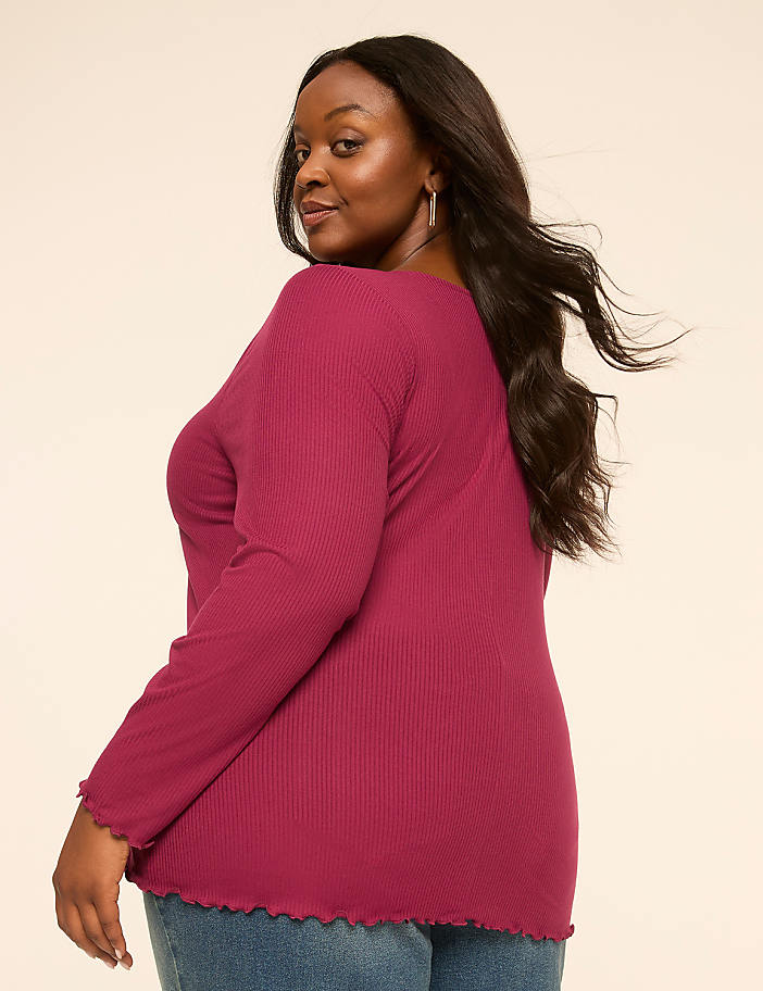 A woman in a magenta ribbed top and blue jeans poses against a plain, light-colored background.