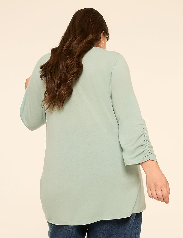 A woman with long, dark hair wearing a light green top with ruched sleeves stands against a plain, light beige background.
