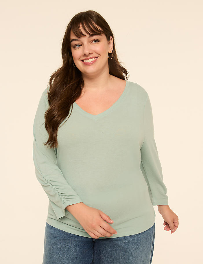 A smiling woman in a light green v-neck shirt and blue jeans stands against a plain, light beige background.