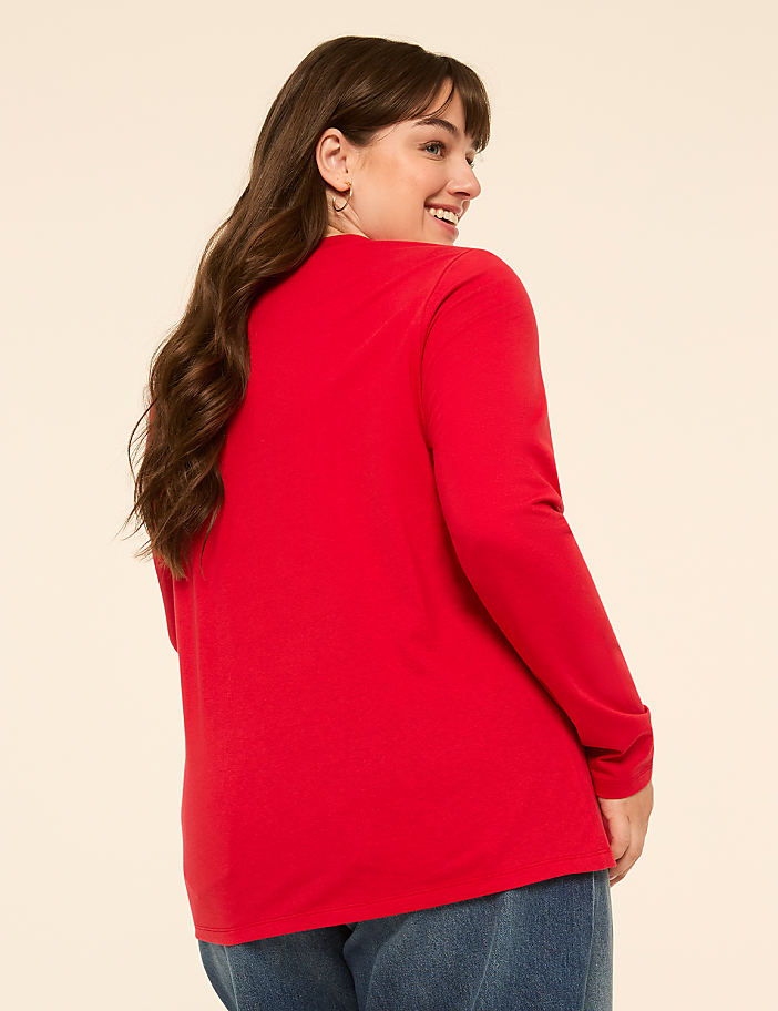 A woman in a red shirt and blue jeans smiles over her shoulder against a plain, light background.