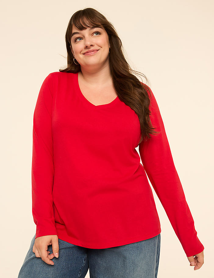 A smiling woman in a red long-sleeved shirt and blue jeans stands against a plain, light-colored background.