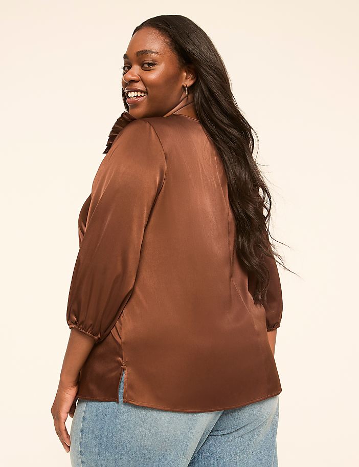A smiling woman in a brown satin blouse and blue jeans stands against a plain, light-colored background.