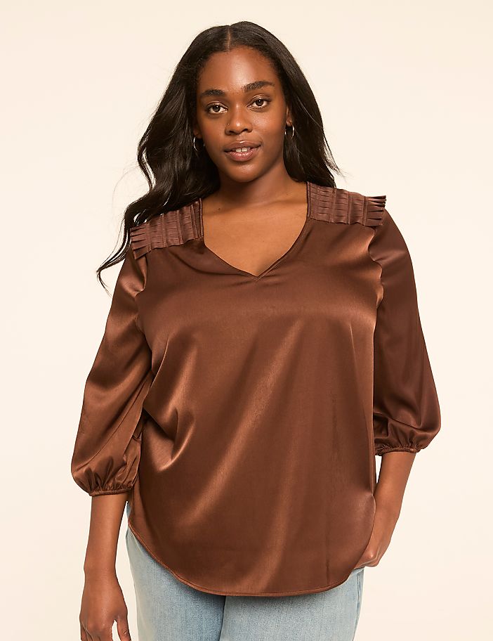 A woman with dark hair and brown eyes, wearing a satin brown blouse with gathered shoulders and light blue jeans, stands against a plain, light-colored background.