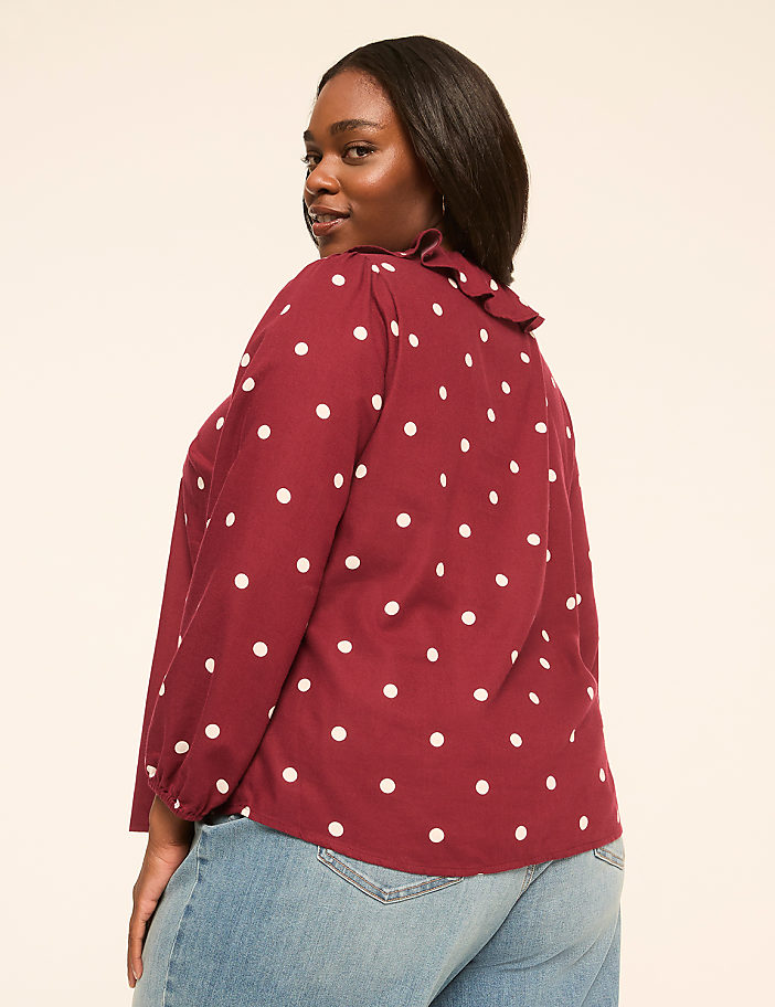 A woman in a maroon polka dot blouse and jeans stands against a plain, light-colored background.