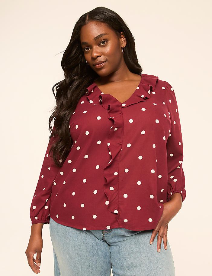 A woman with long dark hair and a polka dot blouse stands against a plain, light-colored background.