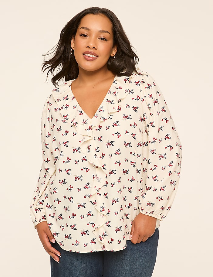 A woman with dark hair and a floral blouse stands against a plain, light-colored background.