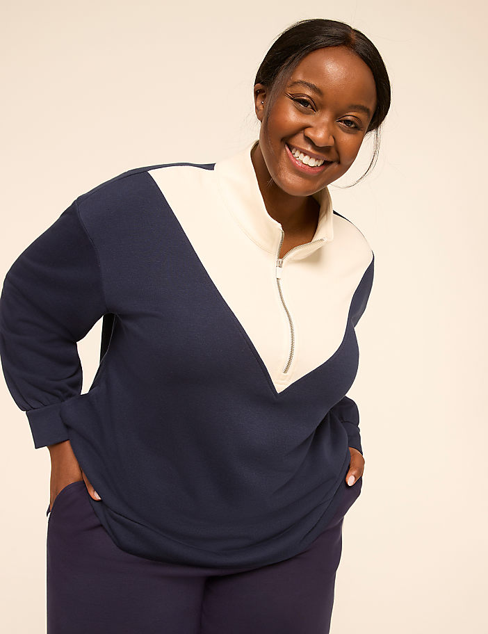 A smiling woman in a navy and cream color-blocked sweatshirt stands against a plain, light-colored background.