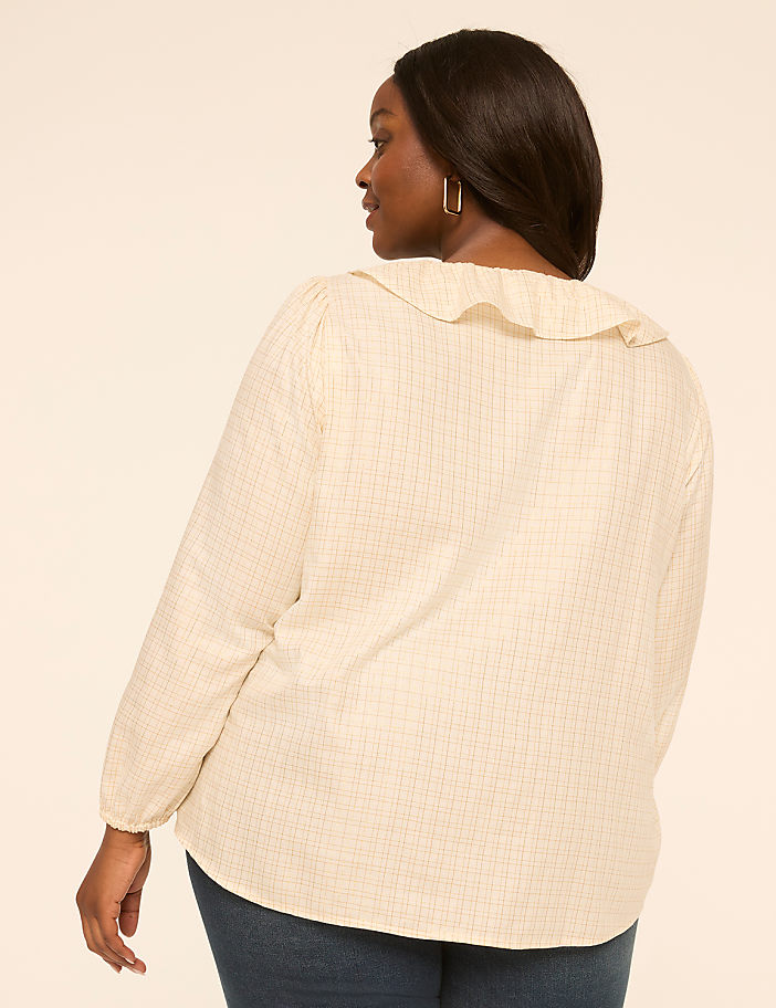 A woman in a cream-colored ruffled blouse and dark jeans stands against a plain, light-colored background.