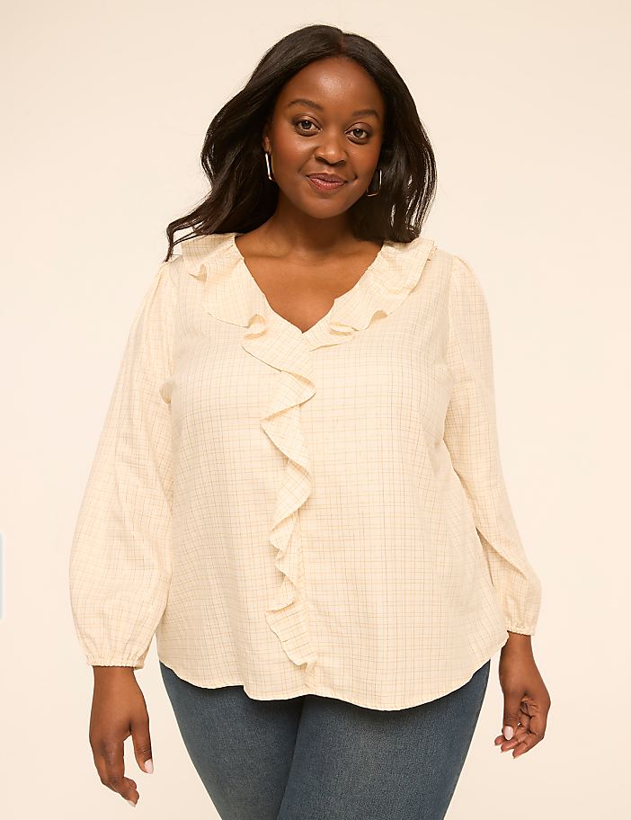 A woman with dark curly hair and a warm smile poses in a cream-colored ruffled blouse and blue jeans against a plain, light-colored background.