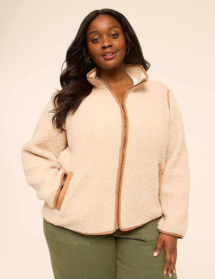A woman in a cream-colored sherpa jacket stands against a plain, light background.