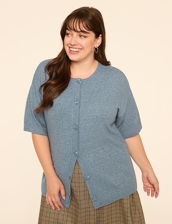 A smiling woman in a blue cardigan and plaid skirt stands against a plain, light-colored background.