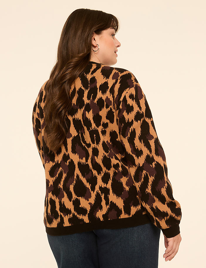 A woman with long brown hair wearing a leopard print sweater and blue jeans stands against a plain, light-colored background.