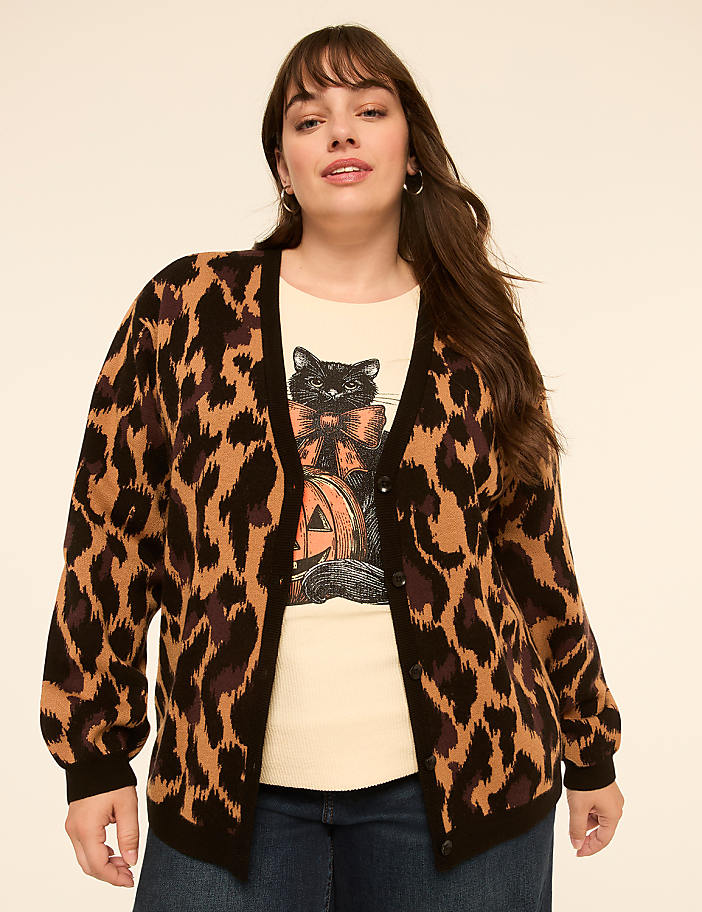 A woman in a leopard print cardigan and a t-shirt with a black cat and pumpkin stands against a plain, light-colored background.